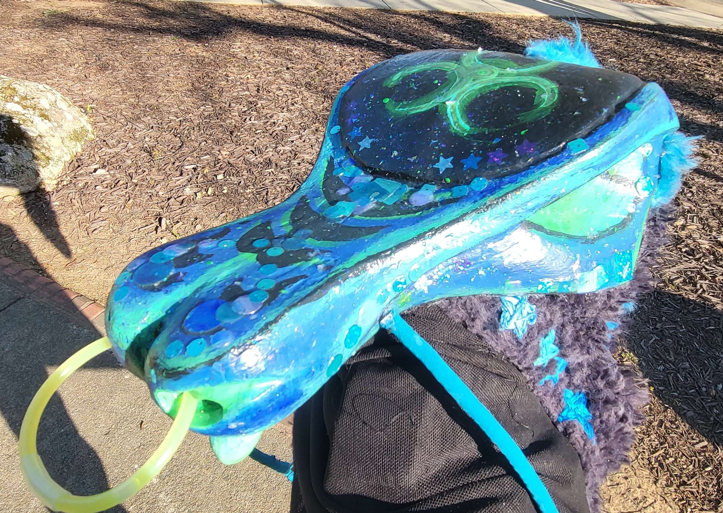 A top 3/4 image of the weevil head, showing the patterning, biohazard sign, sequins, stickers, and glitter. It is colored in various shades of black, blue, and green.